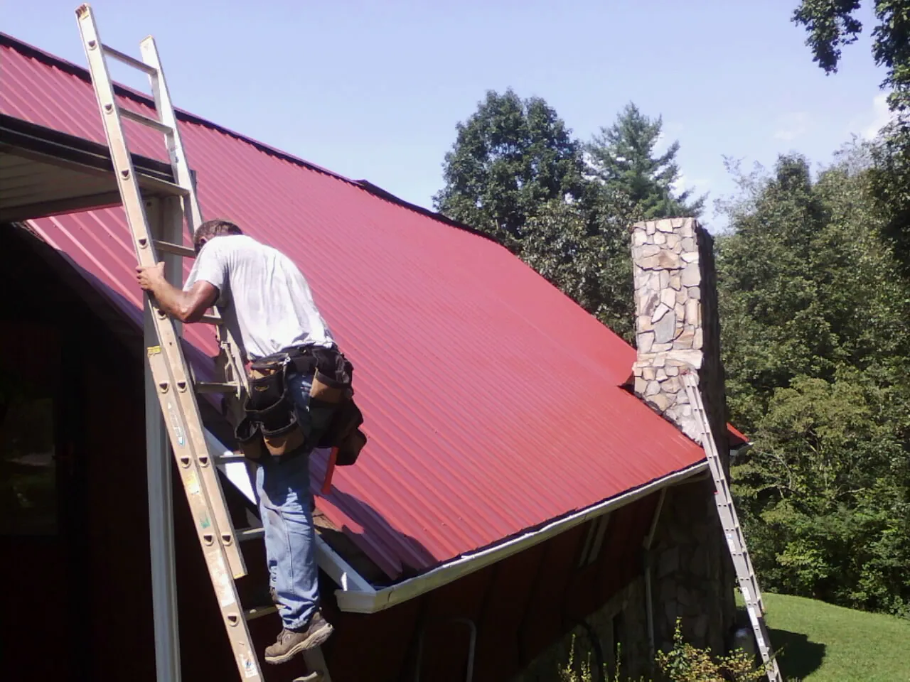 Roofer on a ladder beside a red metal roof and stone chimney — Soffit & Fascia Repair by Roofing Brothers in Sugarloaf